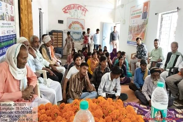 कमरे में कुर्सियों और फर्श पर बैठे बुजुर्ग, बच्चे, युवा और ग्रामीण, सामने मेज पर गेंदे के फूल रखे हैं