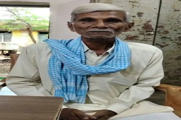 कुर्सी पर बैठे सफेद कपड़े और नीला चेकदार गमछा पहने बुजुर्ग व्यक्ति