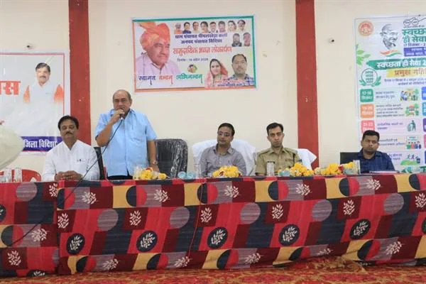 मंच पर मेज के पीछे बैठे अधिकारियों के बीच माइक से संबोधित करते एक व्यक्ति