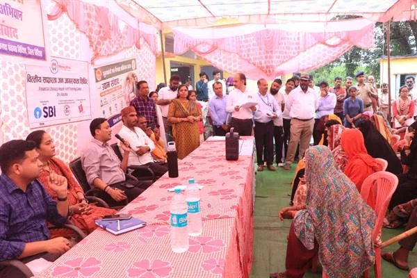 गुलाबी शामियाने के नीचे मेज के पास खड़े अधिकारी सामने कुर्सियों पर बैठी ग्रामीण महिलाओं से संवाद करते हुए