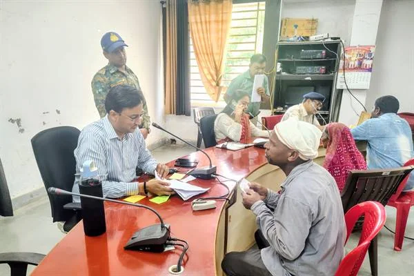 कार्यालय के सभा कक्ष में मेज के सामने बैठे आवेदक की बात सुनते अधिकारी और पीछे मौजूद अन्य लोग