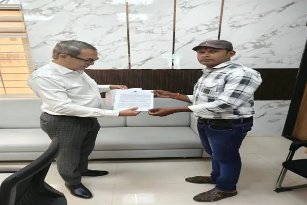 कार्यालय में सफेद शर्ट पहने एक अधिकारी खड़े होकर टोपी और चेक शर्ट पहने एक युवक को कागज का आदेश-पत्र सौंप रहे हैं; पीछे सोफा और संगमरमर की दीवार दिख रही है।