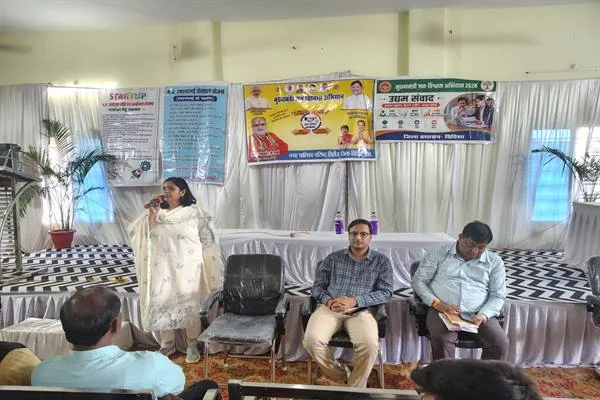 मंच पर खड़ी एक महिला माइक पर बोलती हुईं, बगल में कुर्सियों पर बैठे दो अधिकारी, पीछे उद्यम संवाद और जन विश्वास अभियान के बैनर
